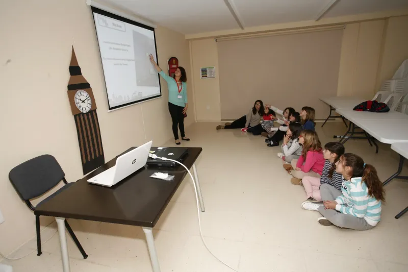 Profesora proyecta una presentación a alumnos sentados en el suelo junto a un Big Ben decorativo