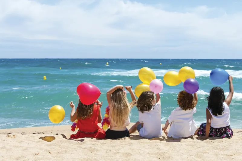 Niños sentados de espaldas en la orilla sujetan globos de colores frente al mar