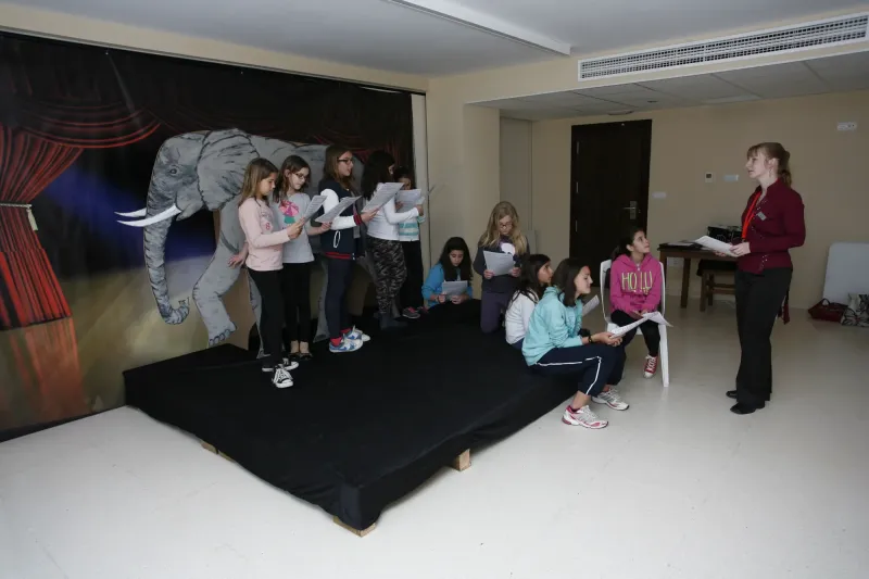 Alumnas ensayan junto al decorado de elefante mientras otras esperan sentadas